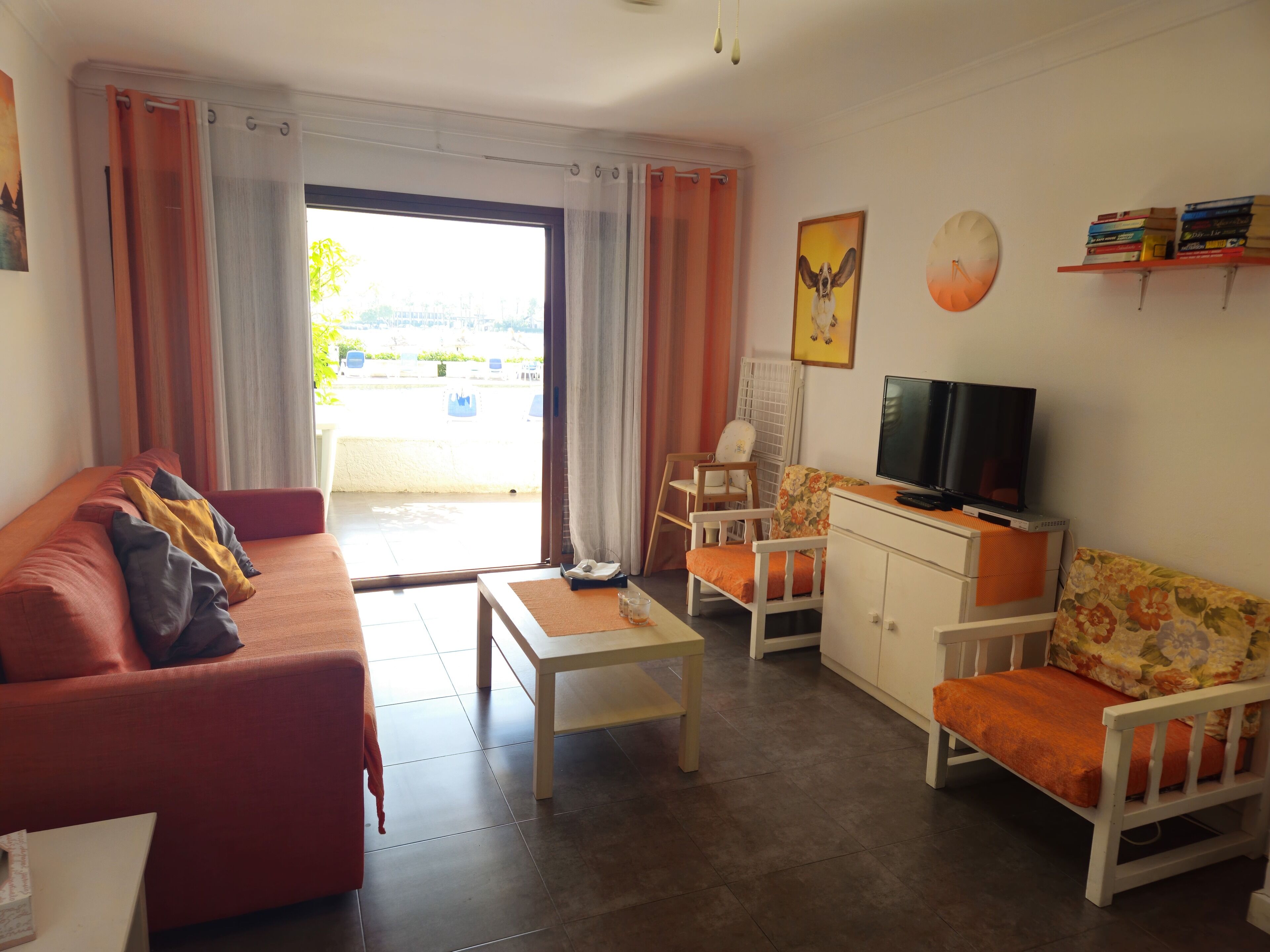 A living room with a sofa, a television, and a small table. There is a window with curtains, a chair, and a bookshelf with books.