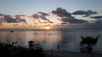 A sunset over a calm sea with boats, a beach, and some vegetation in the foreground.