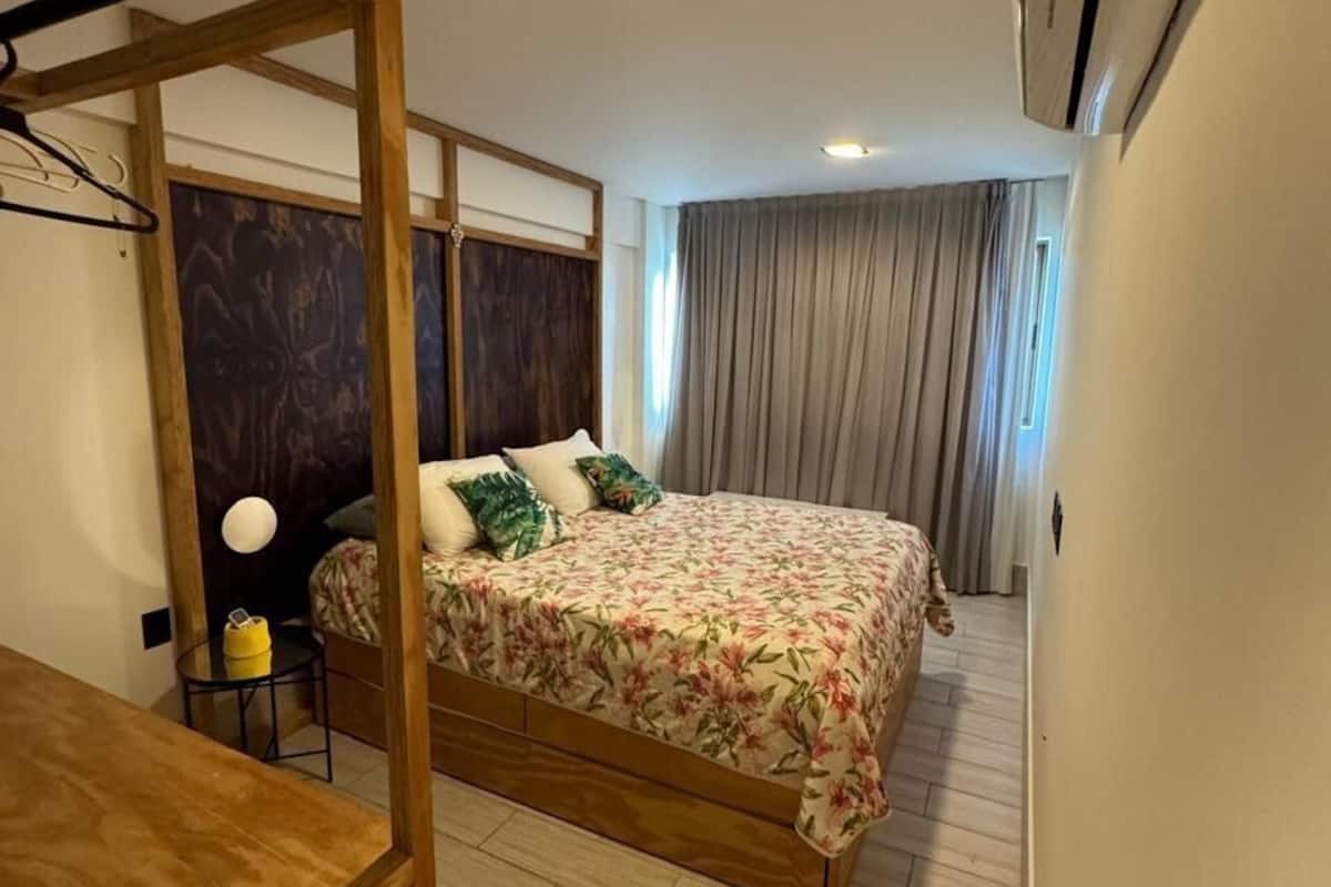 A bedroom with a bed, a wooden desk, a nightstand, a wall-mounted air conditioner, and a window with curtains.