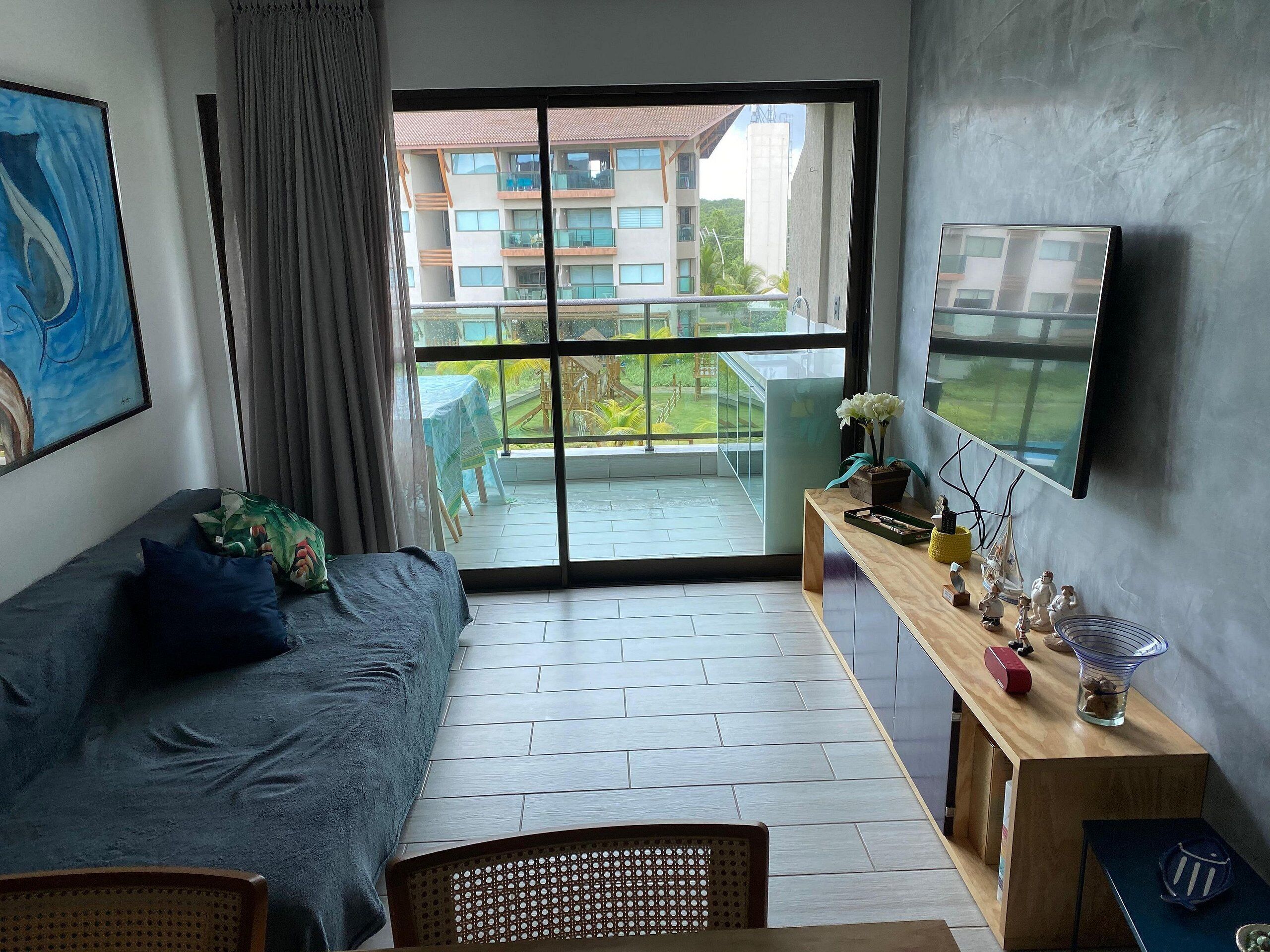 Een kamer met een bed, bureau, stoel, televisie en een balkon met uitzicht op gebouwen en groen.
