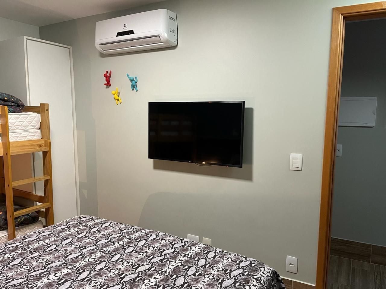 A bedroom with a bunk bed, a flat-screen TV mounted on the wall, and an air conditioning unit above it.