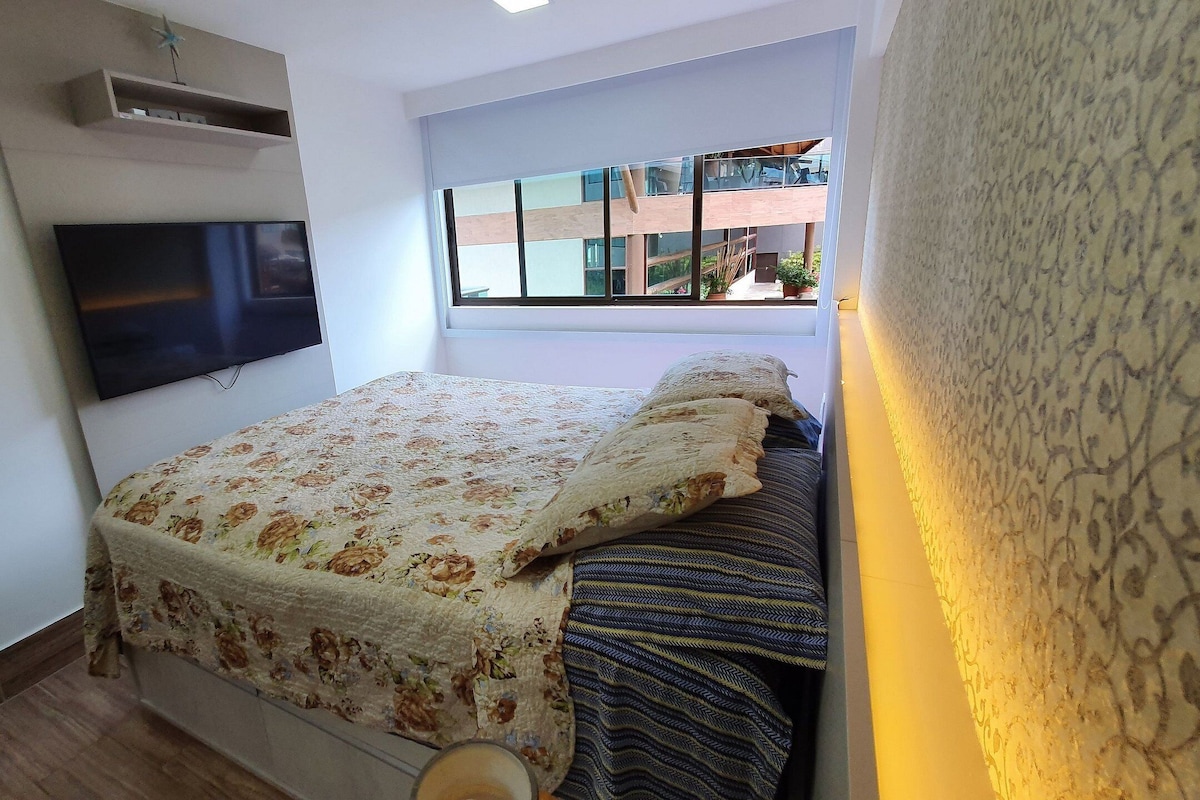 Une chambre à coucher avec un lit, un téléviseur à écran plat fixé au mur et une fenêtre donnant sur des immeubles.