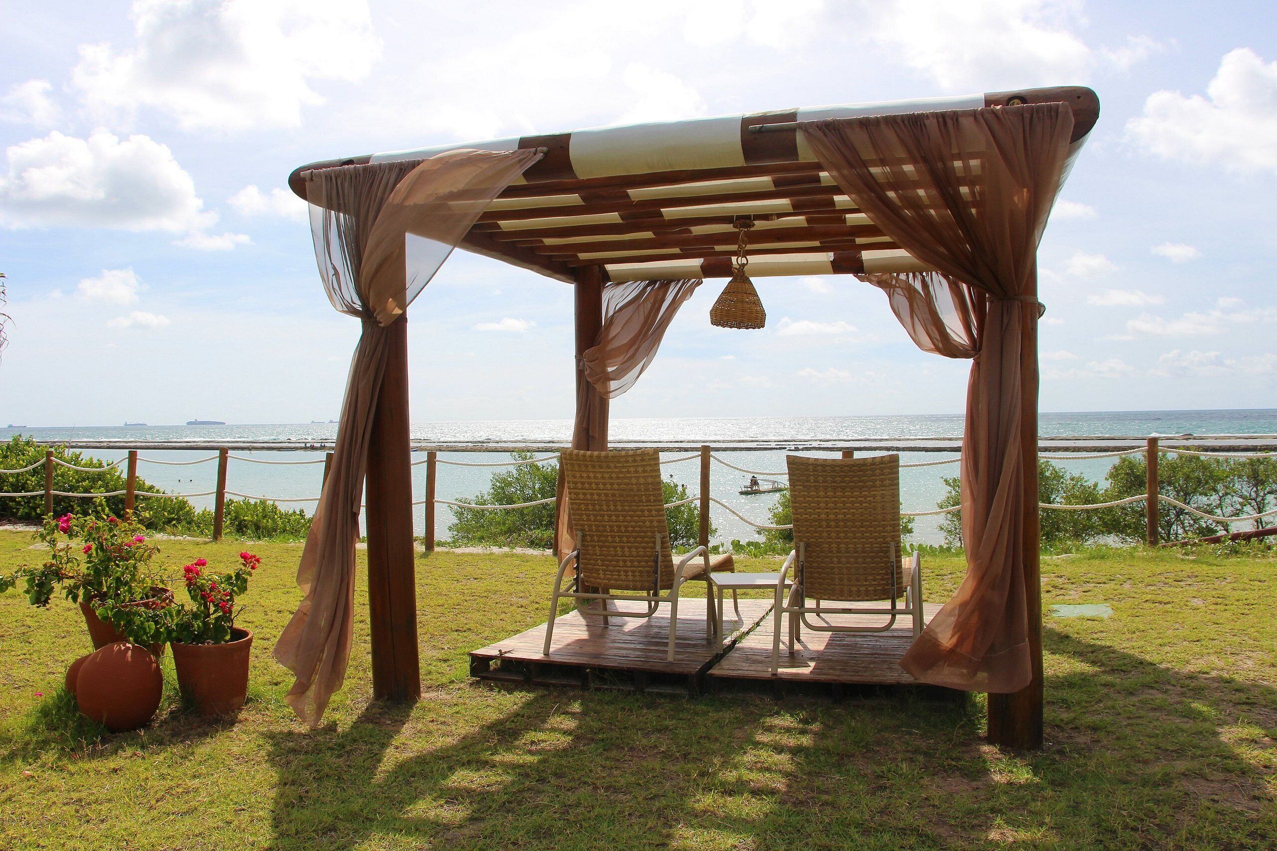 Una pérgola de madera con dos sillas de mimbre y una mesa, con vista a un cuerpo de agua.