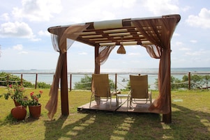 A wooden pergola with two wicker chairs and a table, overlooking a body of water.