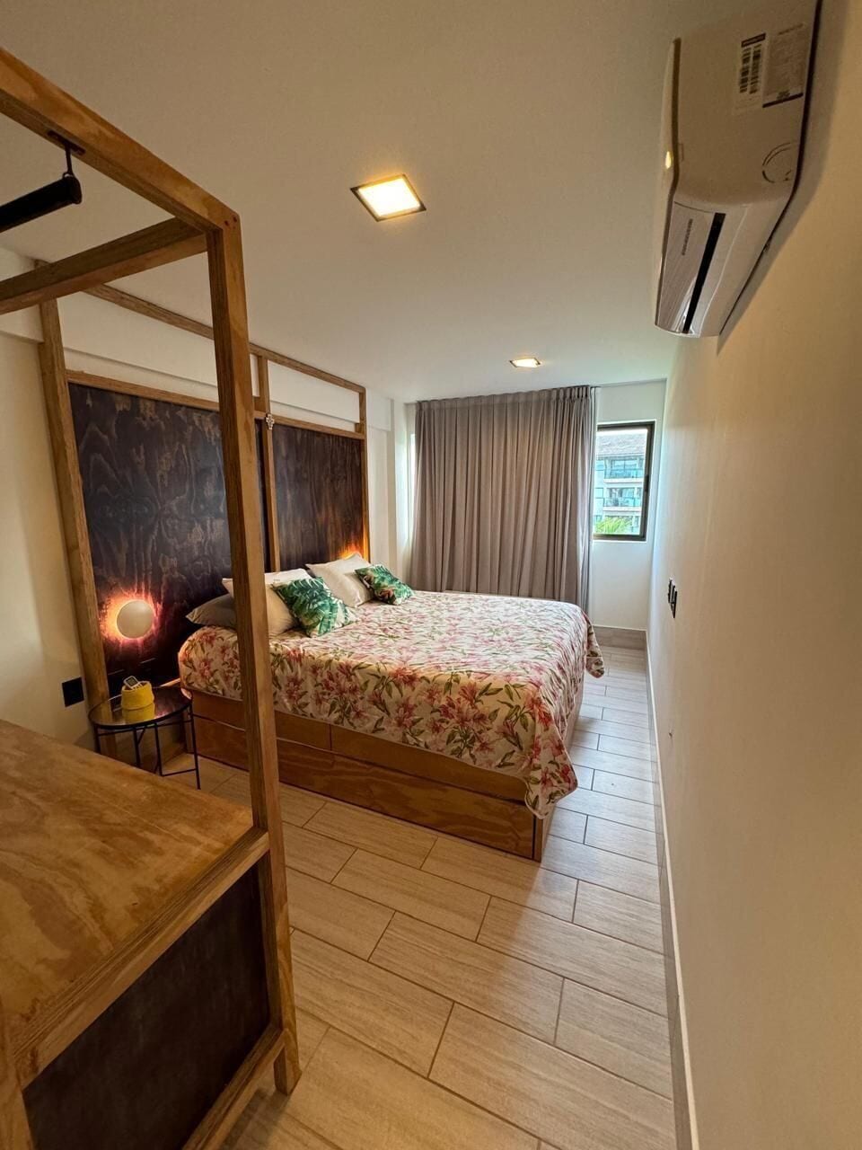 Un dormitorio con estructura de cama de madera, una mesita de noche, aire acondicionado instalado en la pared y una ventana con cortinas.