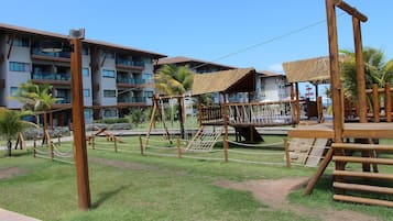 Un parque infantil con estructuras de madera, una palmera y un edificio con balcones al fondo.