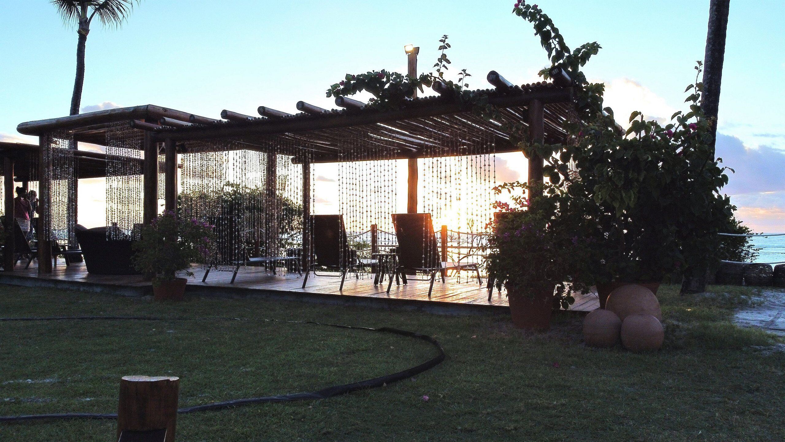 A covered outdoor seating area with chairs and tables, surrounded by greenery and a view of the sea.