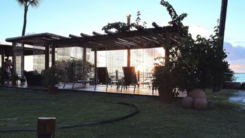 A covered outdoor seating area with chairs and tables, surrounded by greenery and a view of the sea.