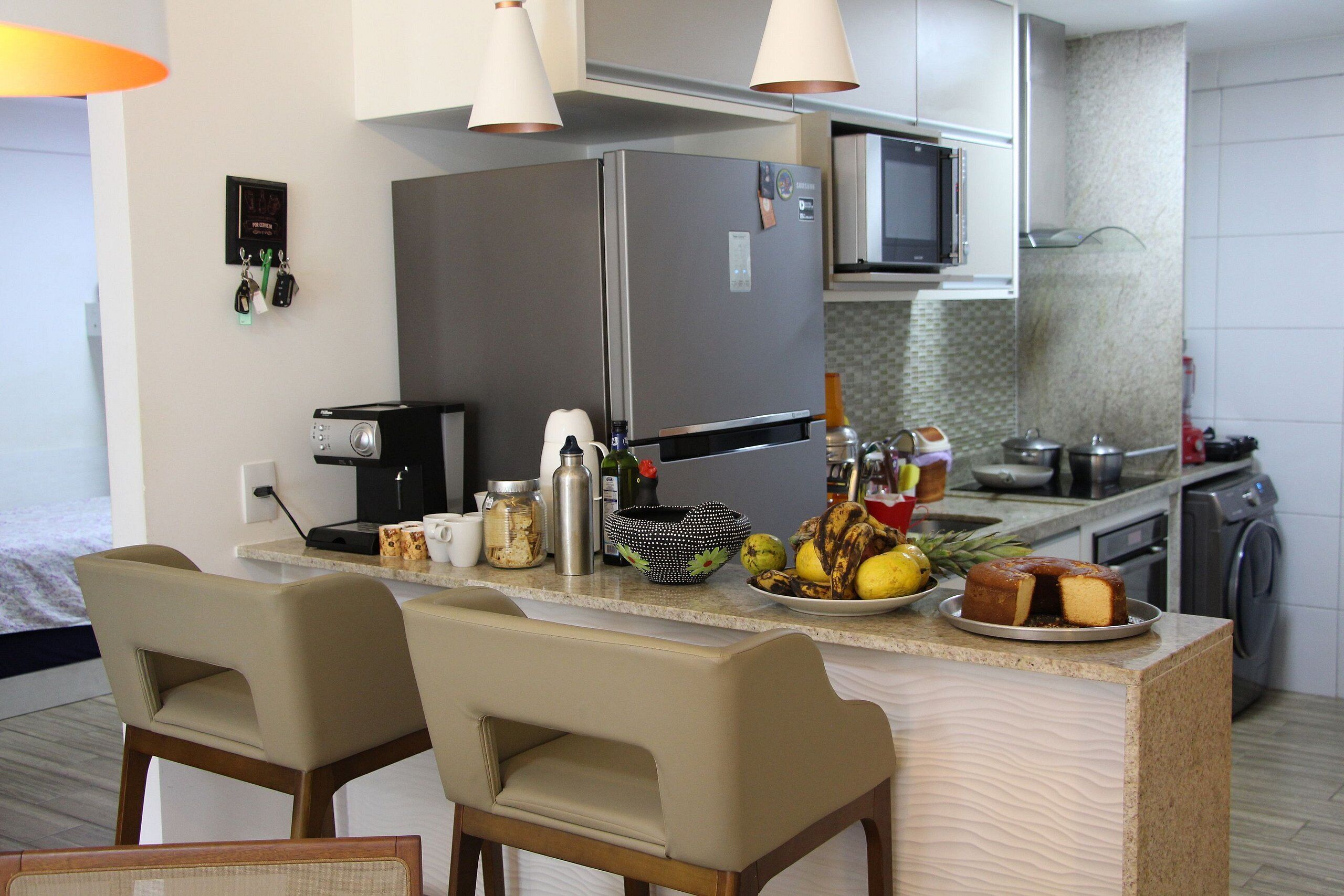 A modern kitchen with a refrigerator, microwave, and coffee maker. There are two bar stools and a fruit bowl on the counter.