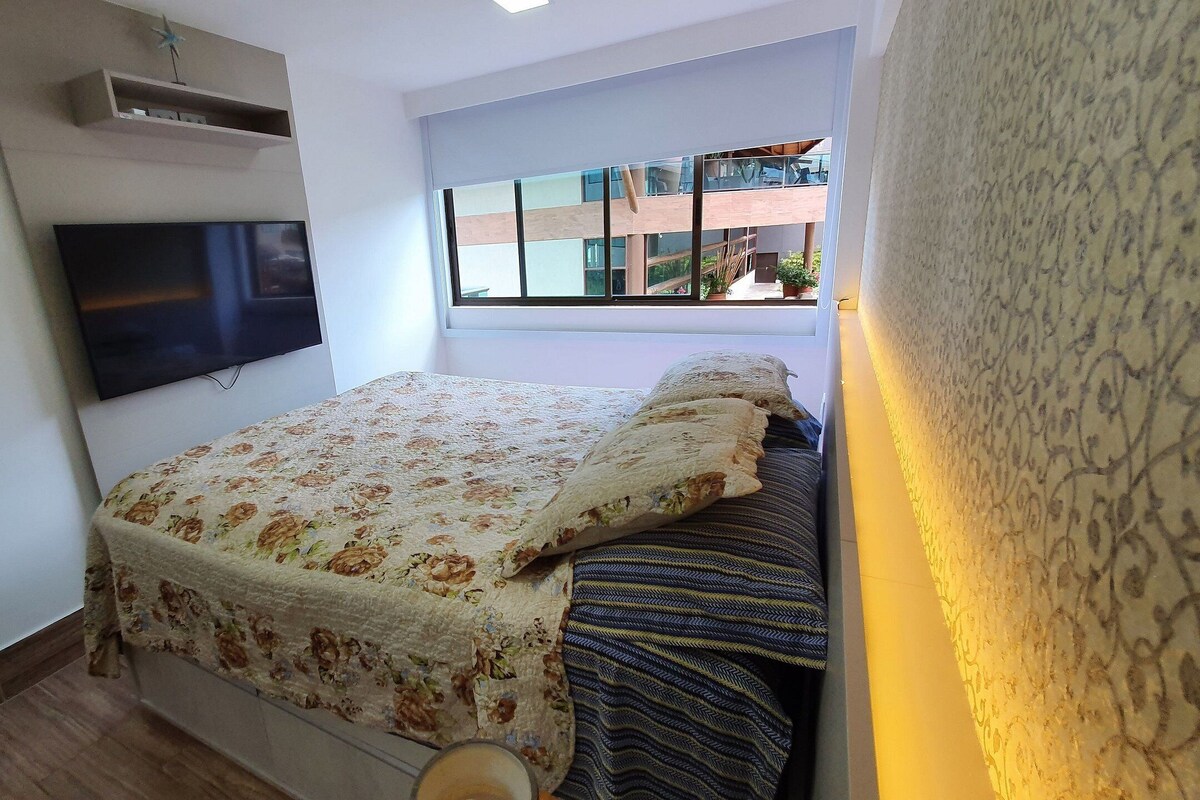 A bedroom with a bed, a flat-screen TV mounted on the wall, and a window with a view of buildings.