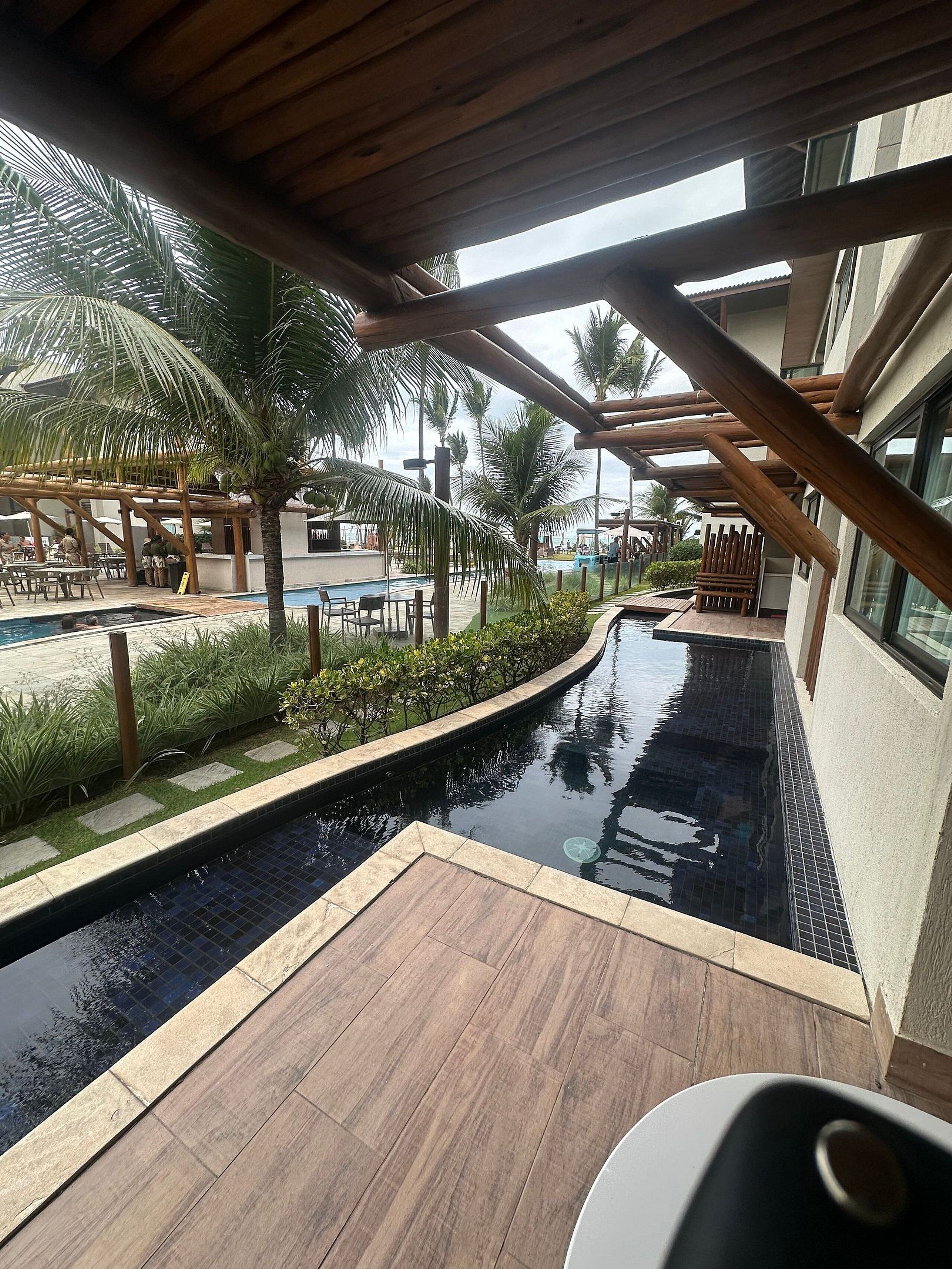 Un espace piscine doté d’une terrasse en bois, de palmiers et d’un coin salon abrité.
