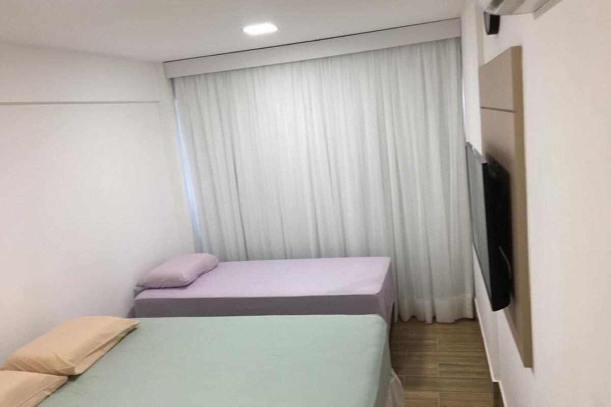 A small, modern room with two beds, a flat-screen TV mounted on the wall, and a window with white curtains.