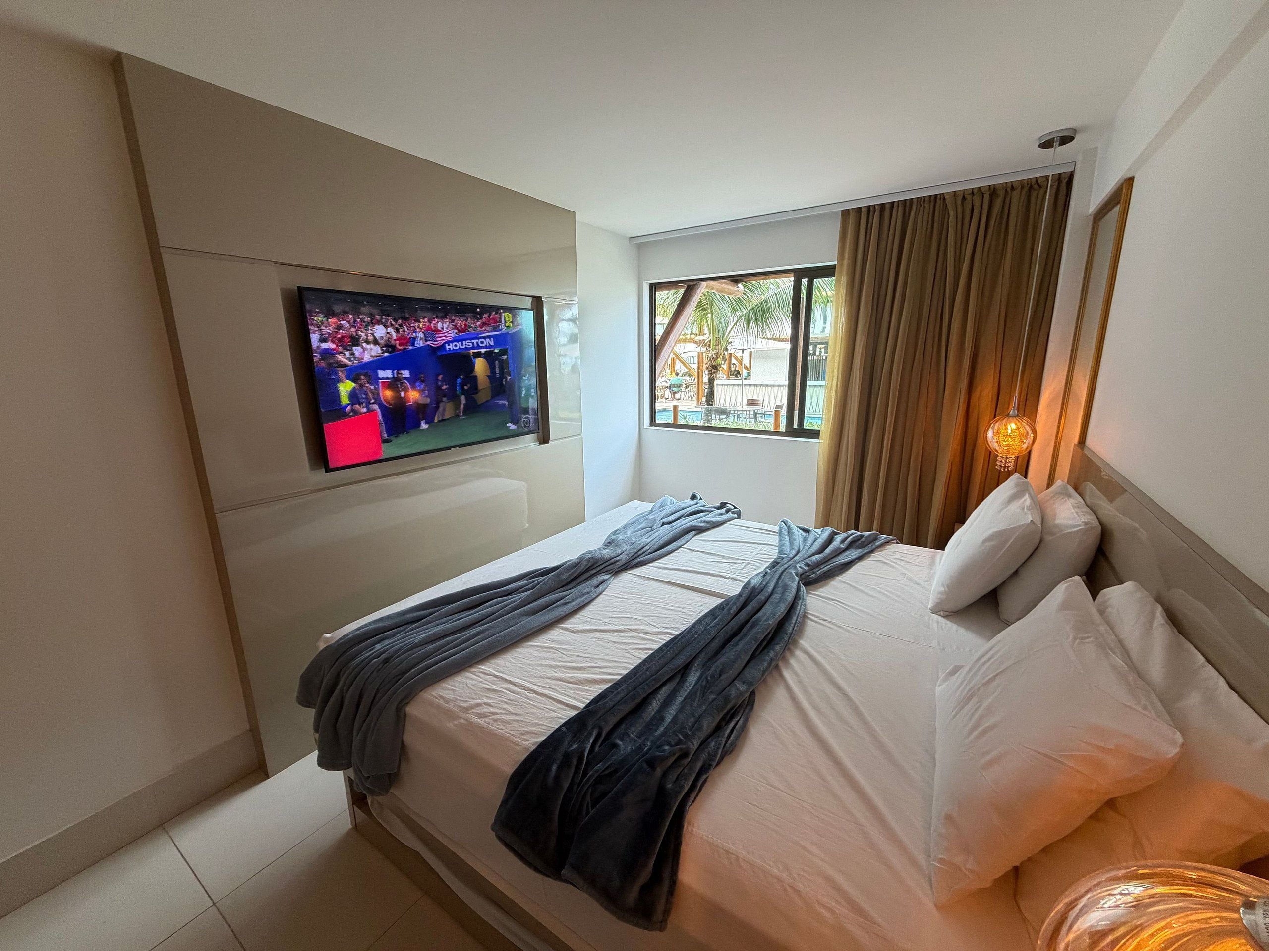 Une chambre d’hôtel avec un lit, un téléviseur fixé au mur, une fenêtre avec des rideaux et une lampe de chevet.