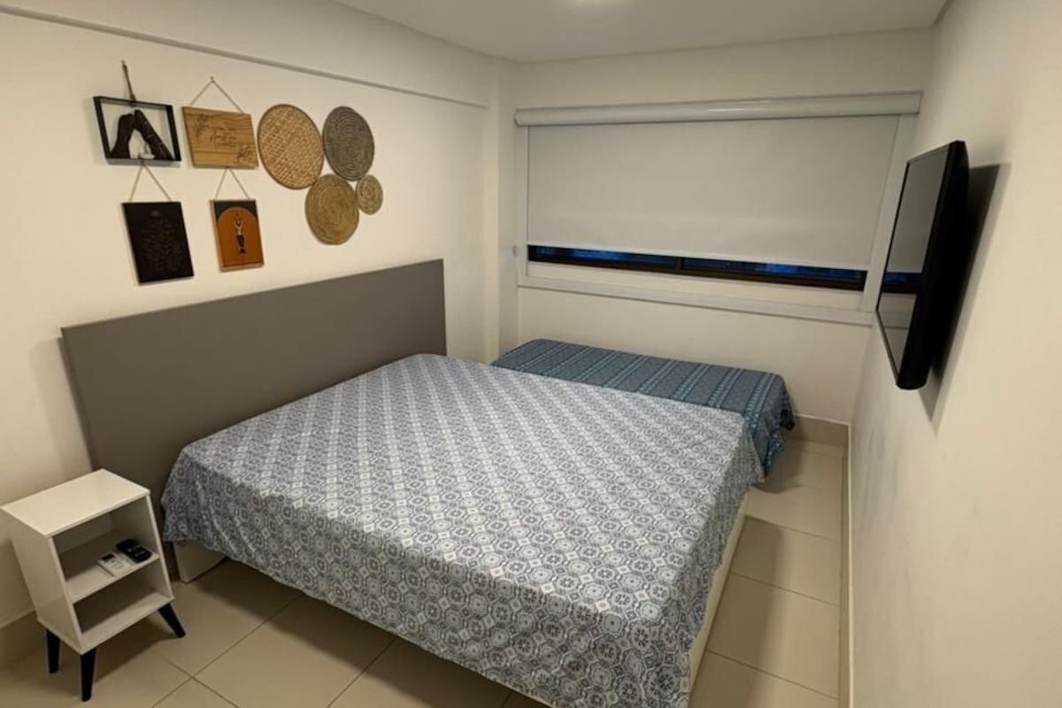 A bedroom with a bed, a bedside table, a wall-mounted TV, and a window with blinds.