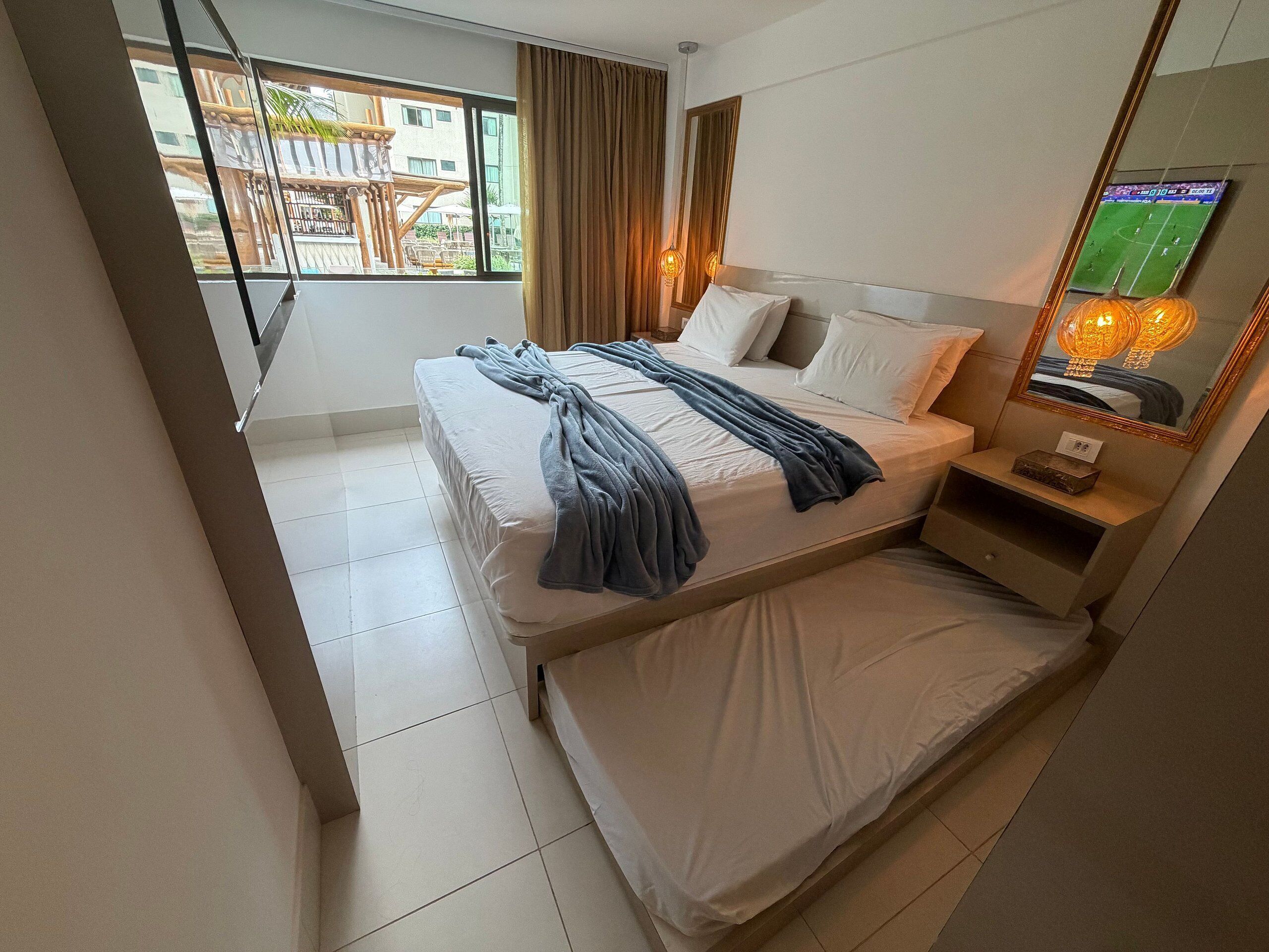 A hotel room with a bed, a bedside table, a mirror, and a window with curtains.