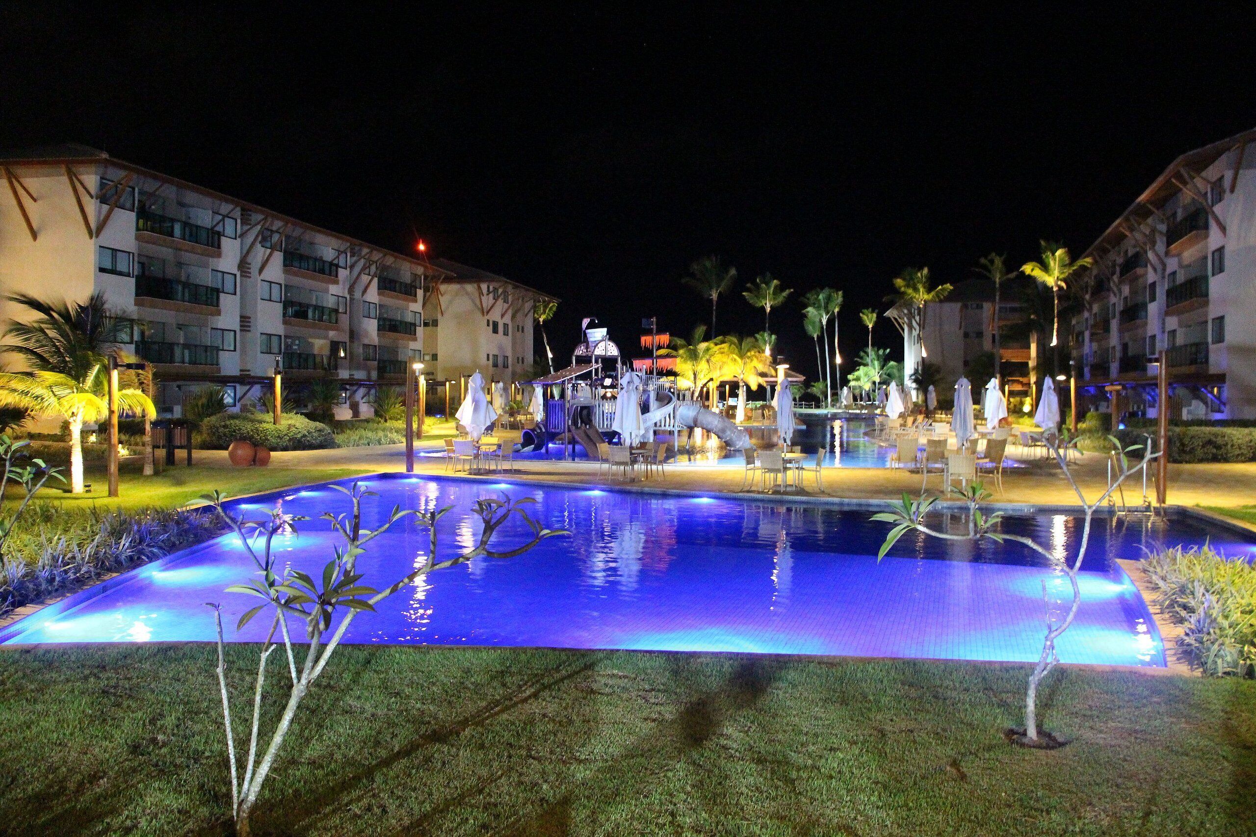 A nighttime resort pool area with a central water feature, surrounded by palm trees and lounge chairs.