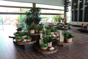 A lobby with multiple wooden tables with potted plants, a sofa, and a large window with a view of outdoor seating. - Polinesia Resort - Porto de Galinhas - Acesso ao Hotel Samoa. (Ipojuca)