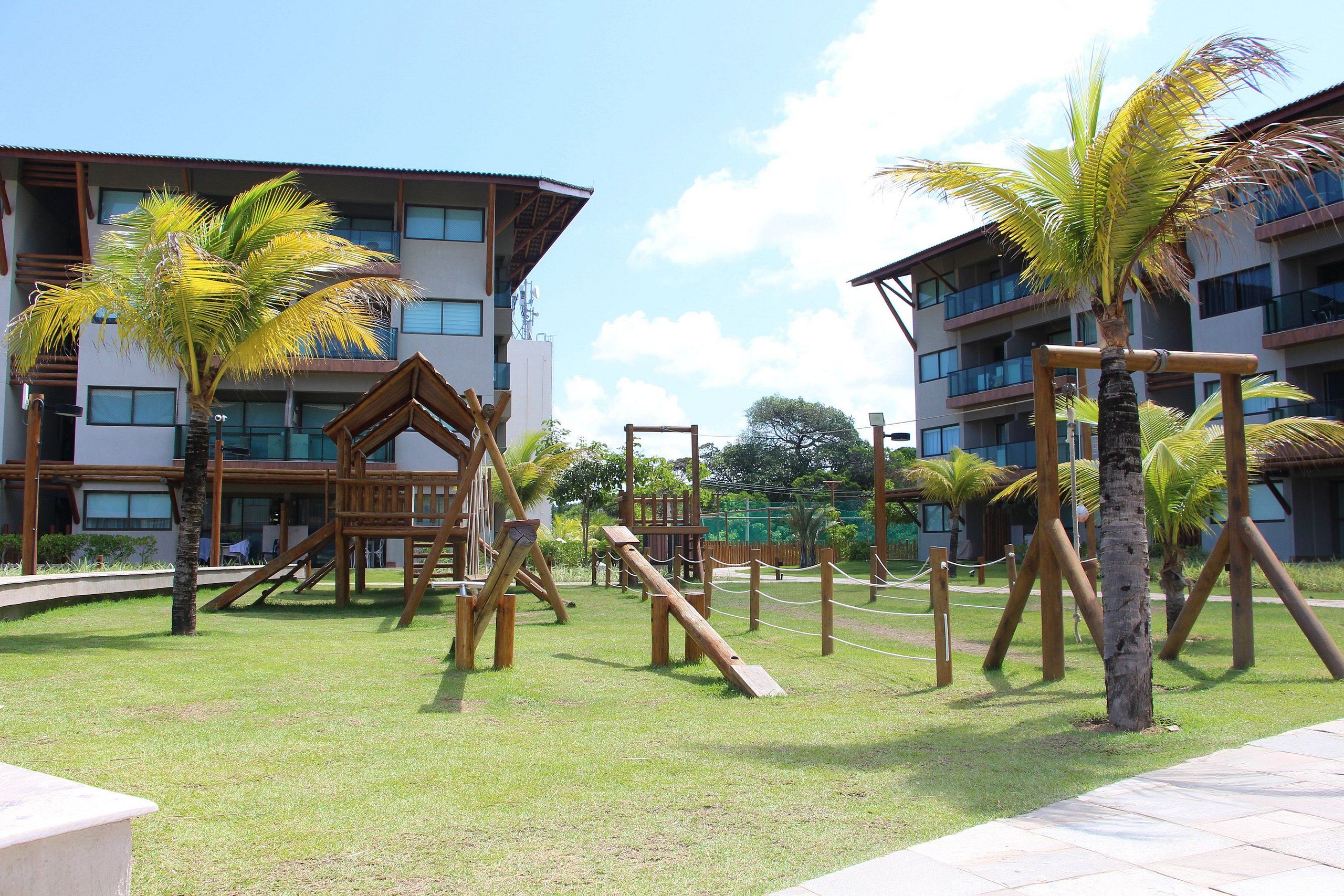 A playground with wooden structures and palm trees, surrounded by buildings with balconies.