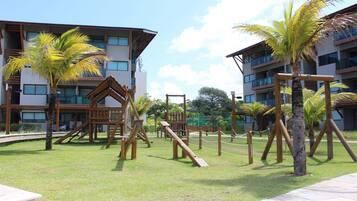 A playground with wooden structures and palm trees, surrounded by buildings with balconies.