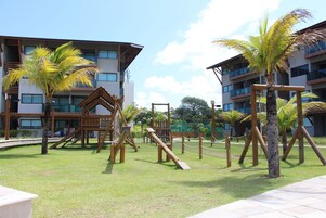 A playground with wooden structures and palm trees, surrounded by buildings with balconies.