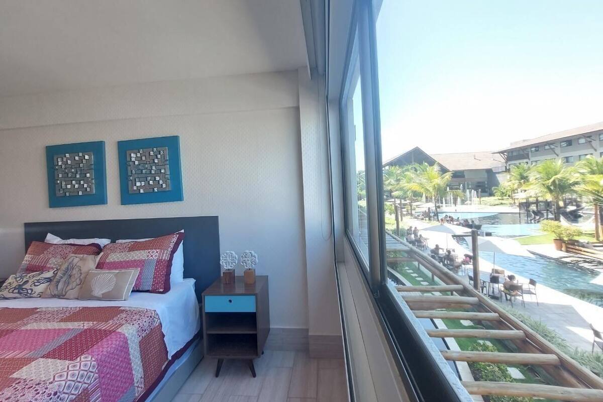 A bedroom with a large bed, a nightstand, and a view of a pool and resort area.