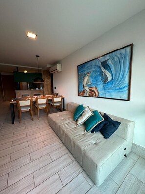 Een moderne woonkamer met een bank, kussens, een eethoek met stoelen, een schilderij aan de muur en een keuken op de achtergrond.