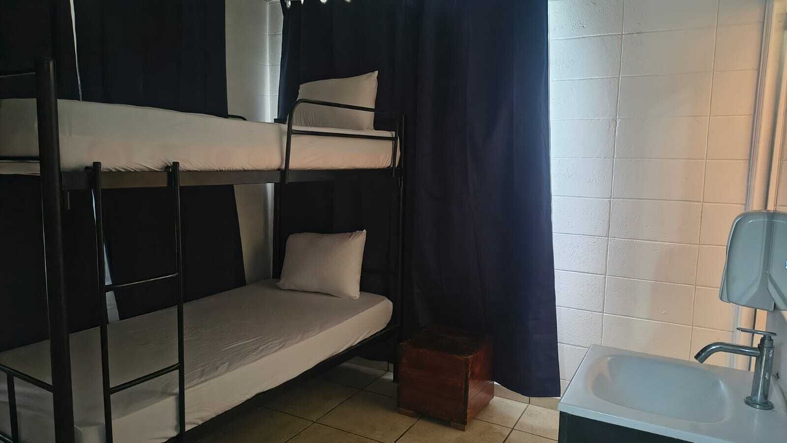 A bunk bed with a small bedside table and a single bed. There is a bathroom with a sink and a mirror.
