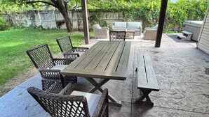 Un patio exterior con una mesa de madera y sillas de mimbre, rodeado por una zona cubierta con luces colgantes.