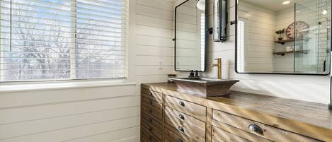 A bathroom with a wooden vanity, a large mirror, and a window with blinds.