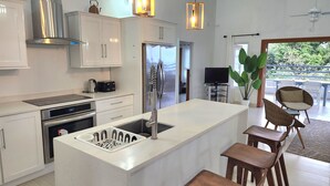Une cuisine moderne avec des meubles blancs, des appareils en acier inoxydable et un îlot central. On y trouve un coin repas avec des chaises et une table en osier, ainsi qu’une grande fenêtre donnant sur un paysage verdoyant.