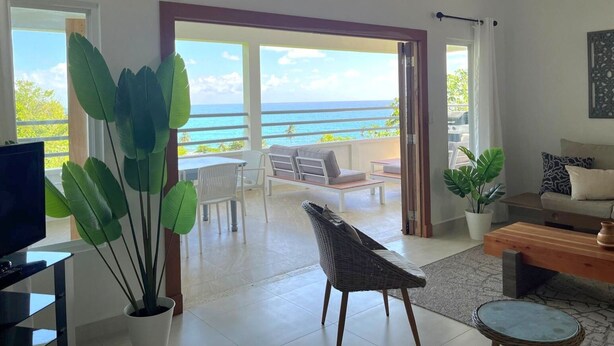 Un vaste espace de vie avec une grande porte-fenêtre coulissante donnant sur un balcon avec vue sur l’océan. On y trouve un téléviseur, un fauteuil en osier et un canapé avec des coussins.