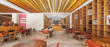 Un interior espacioso con vigas de madera en el techo, una zona de estar central con mesas y sillas redondas, y una estantería llena de objetos decorativos.