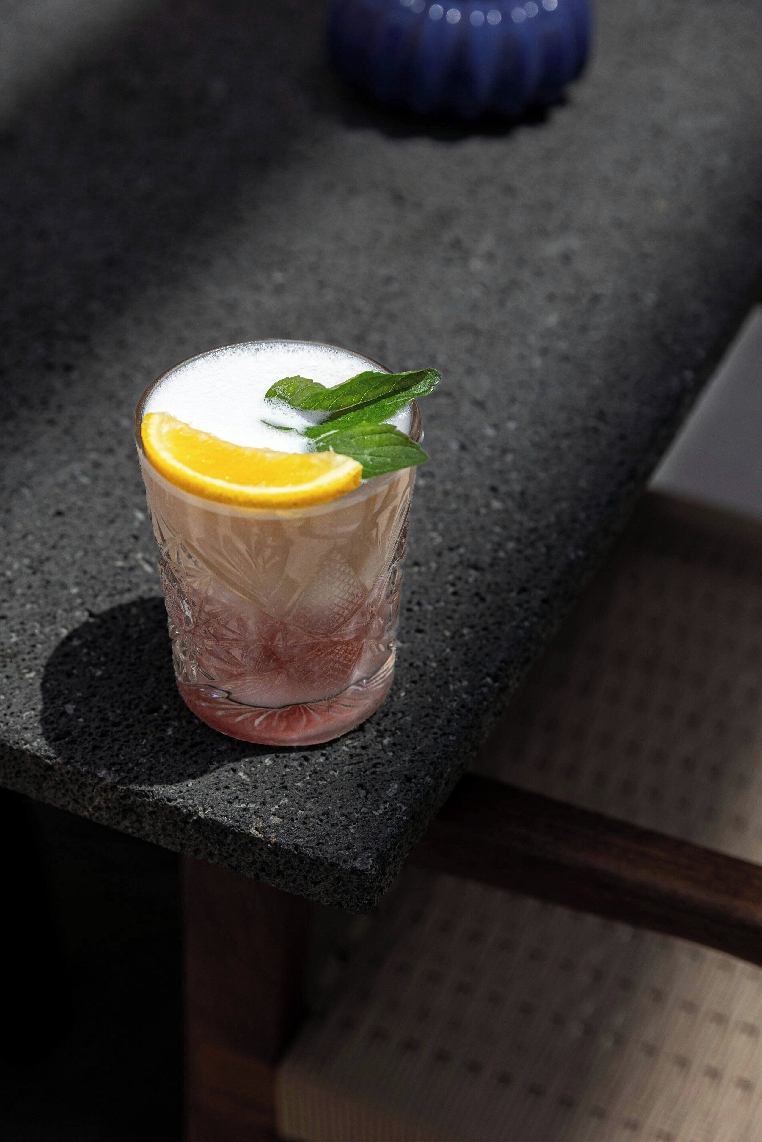 A glass of iced drink with a slice of orange and mint leaves on a dark countertop.