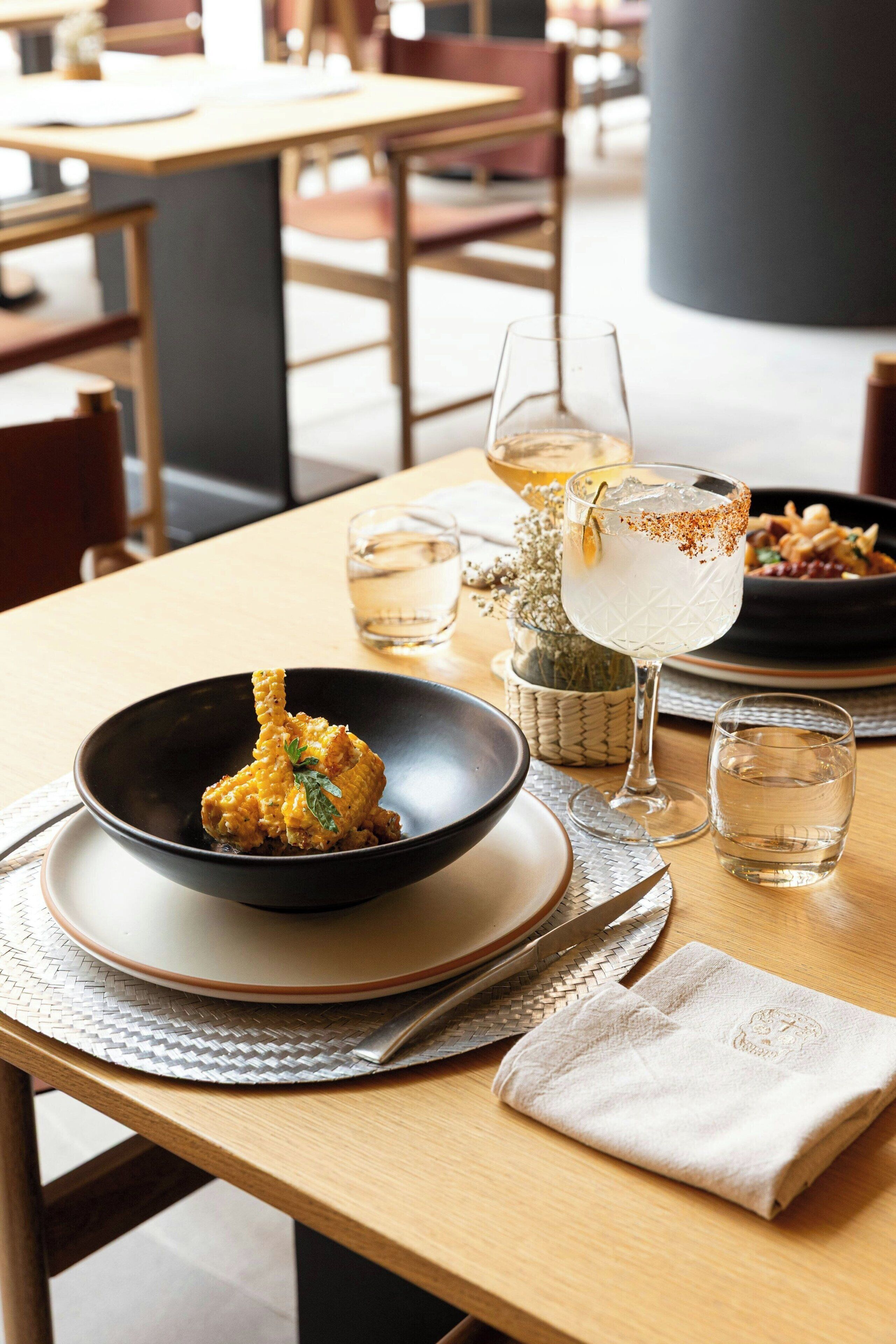 Una mesa de comedor con un tazón negro de comida, una copa de vino blanco y una copa de cóctel con guarnición.