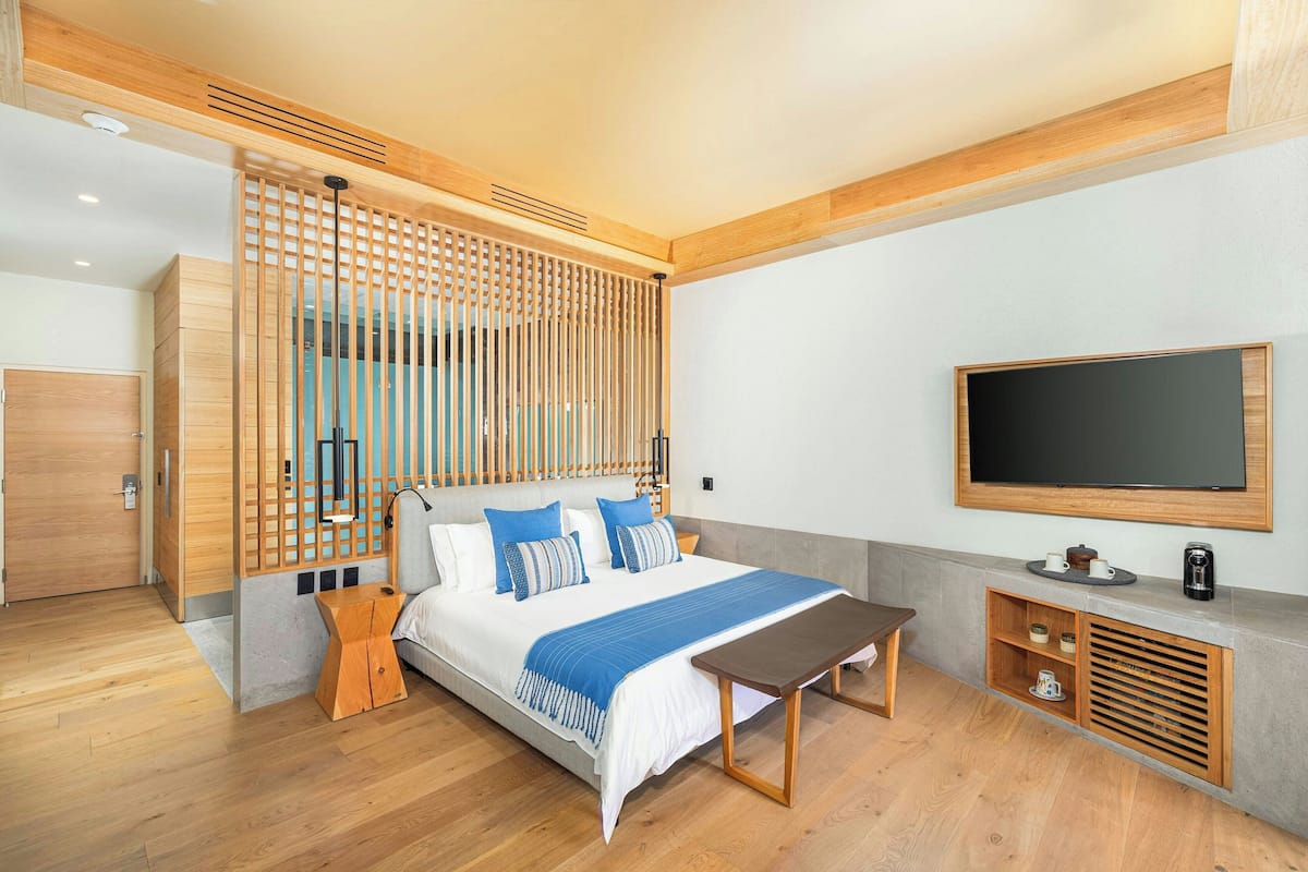 Una habitación de hotel moderna con una cama grande, cabecera de madera, un televisor montado en la pared y una mesita de noche de madera.