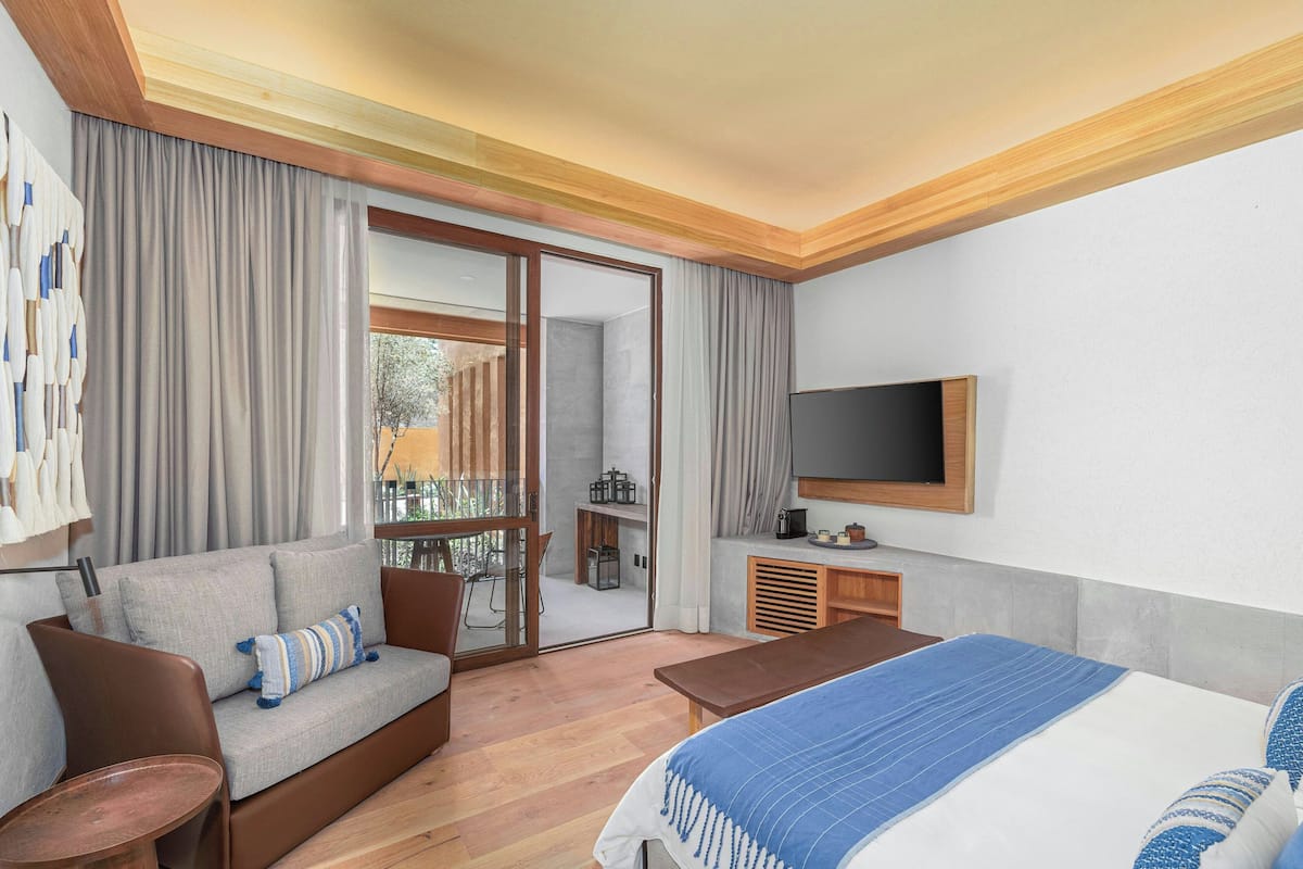Una habitación de hotel moderna con sofá, cama, televisor y una puerta corrediza de vidrio que da a un balcón.