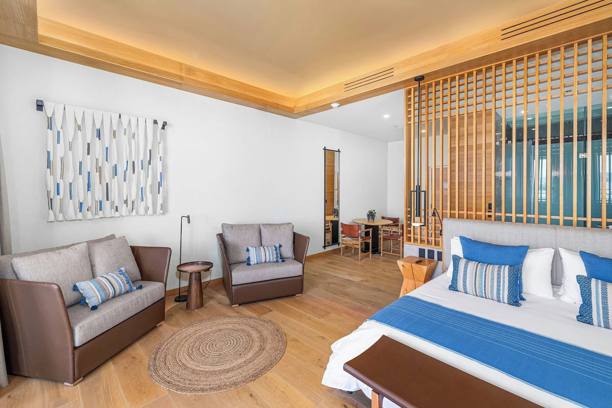 Una habitación de hotel moderna con una cama grande, dos sofás, una mesita redonda y una zona de comedor al fondo.