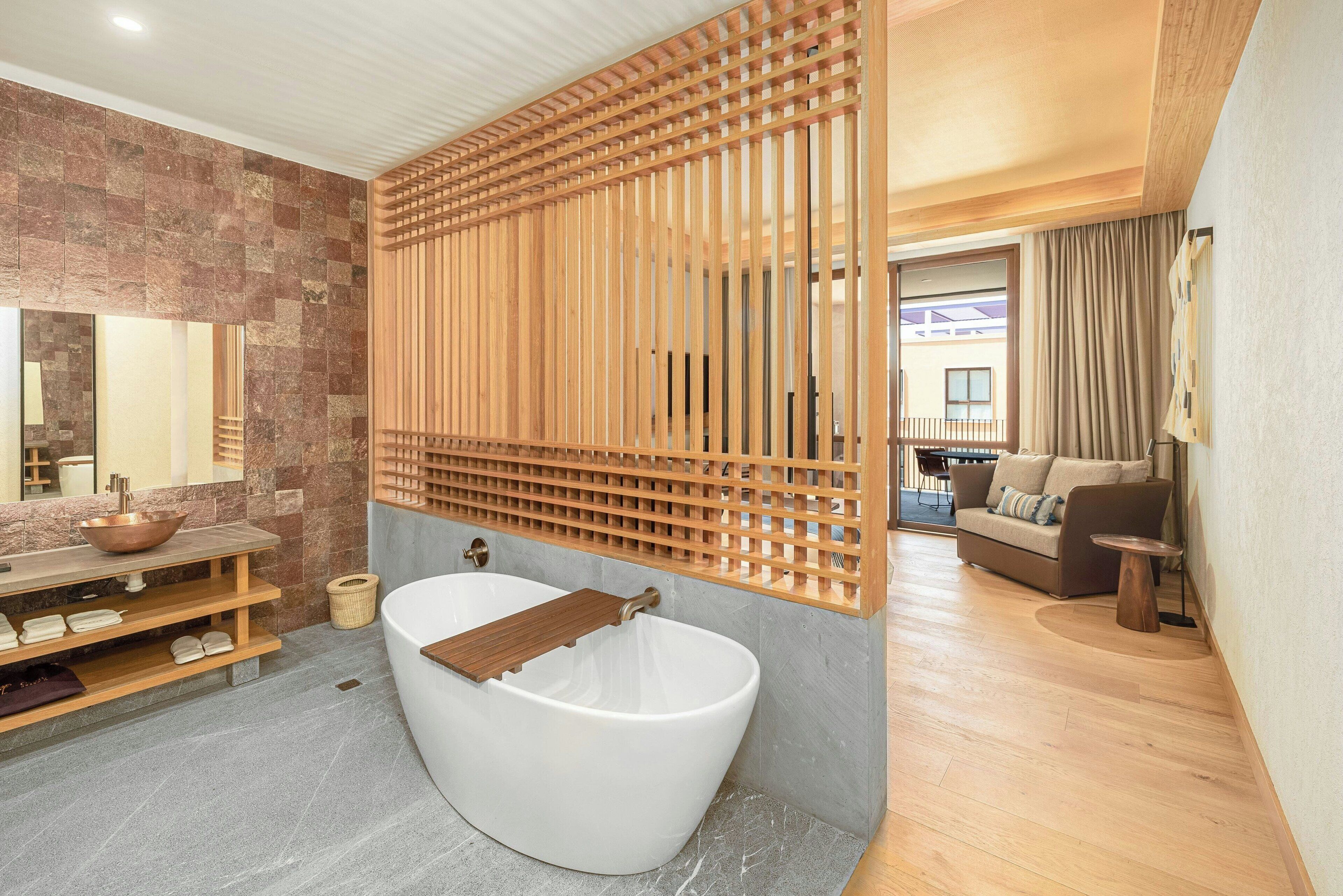 A modern bathroom with a freestanding bathtub, a wooden slatted partition, and a stone wall.