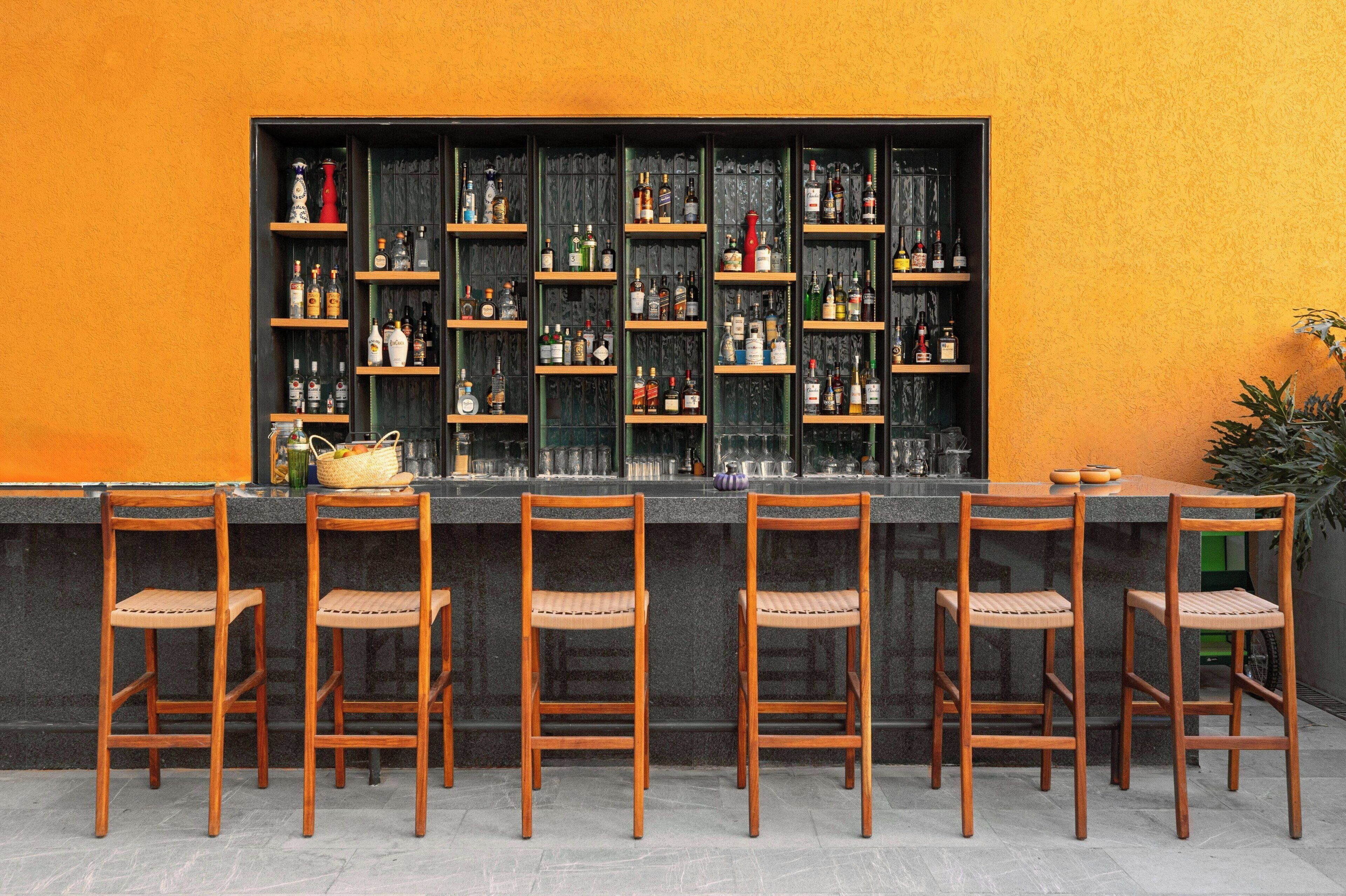 Zona de bar con varias estanterías llenas de botellas, una barra y seis sillas de madera.