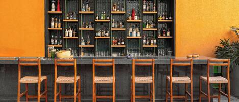 Zona de bar con varias estanterías llenas de botellas, una barra y seis sillas de madera.