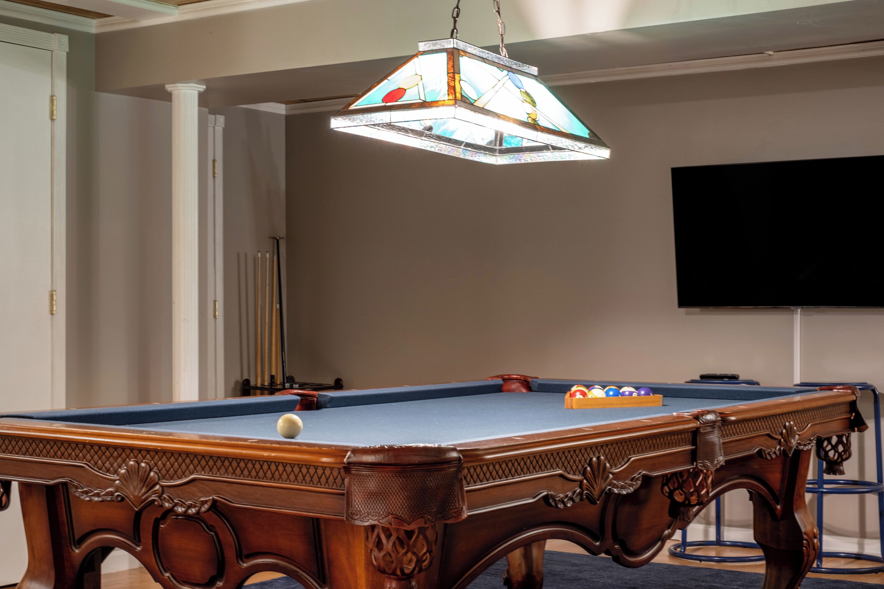 Une table de billard surmontée d’un luminaire en vitrail, un téléviseur fixé au mur et une porte donnant sur une autre pièce.