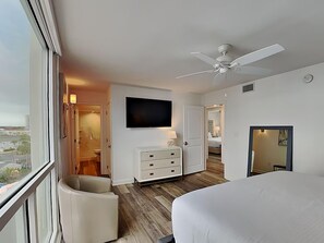A hotel room with a large bed, a ceiling fan, a TV mounted on the wall, a dresser, and a view of the city through the window. - Shoreline Towers 1066 (Destin)