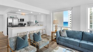 A modern living room with a blue sofa, a dining area with a white island, and a kitchen with stainless steel appliances. - Shoreline Towers 1066 (Destin)