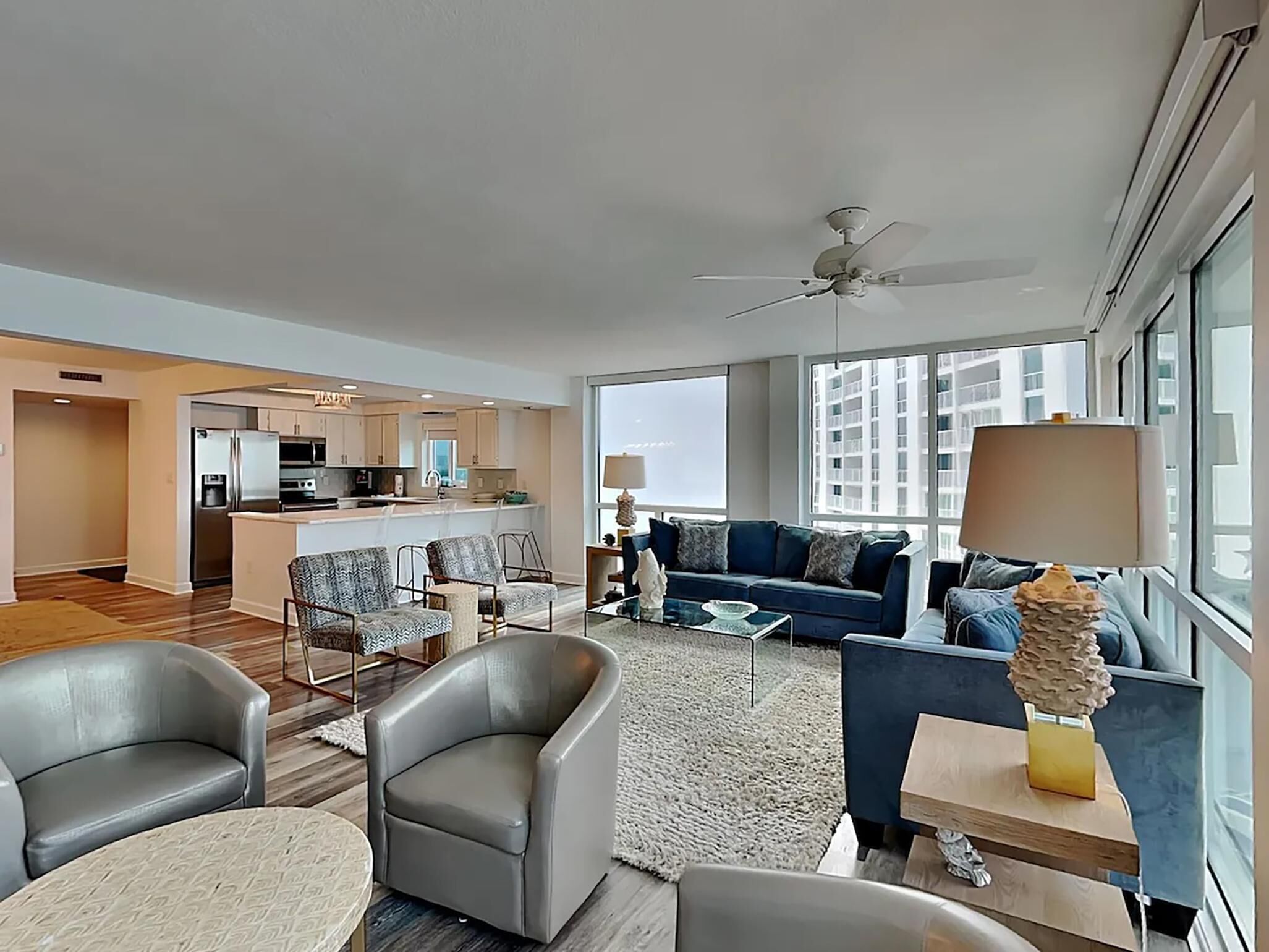 A modern living room with a sofa, armchairs, and a coffee table. There is a kitchen area with a refrigerator and cabinets visible in the background.