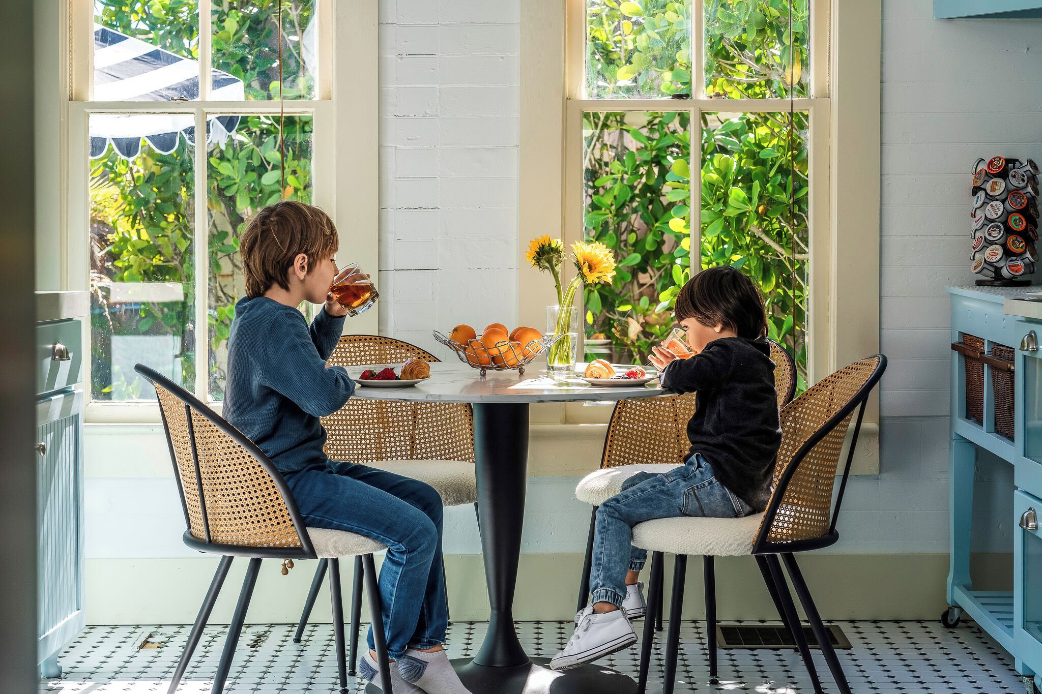 A dining area with wicker chairs, a round table, and a view of greenery through large windows.