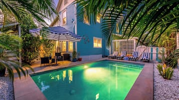 A swimming pool with lounge chairs and umbrellas, surrounded by palm trees and a blue house.