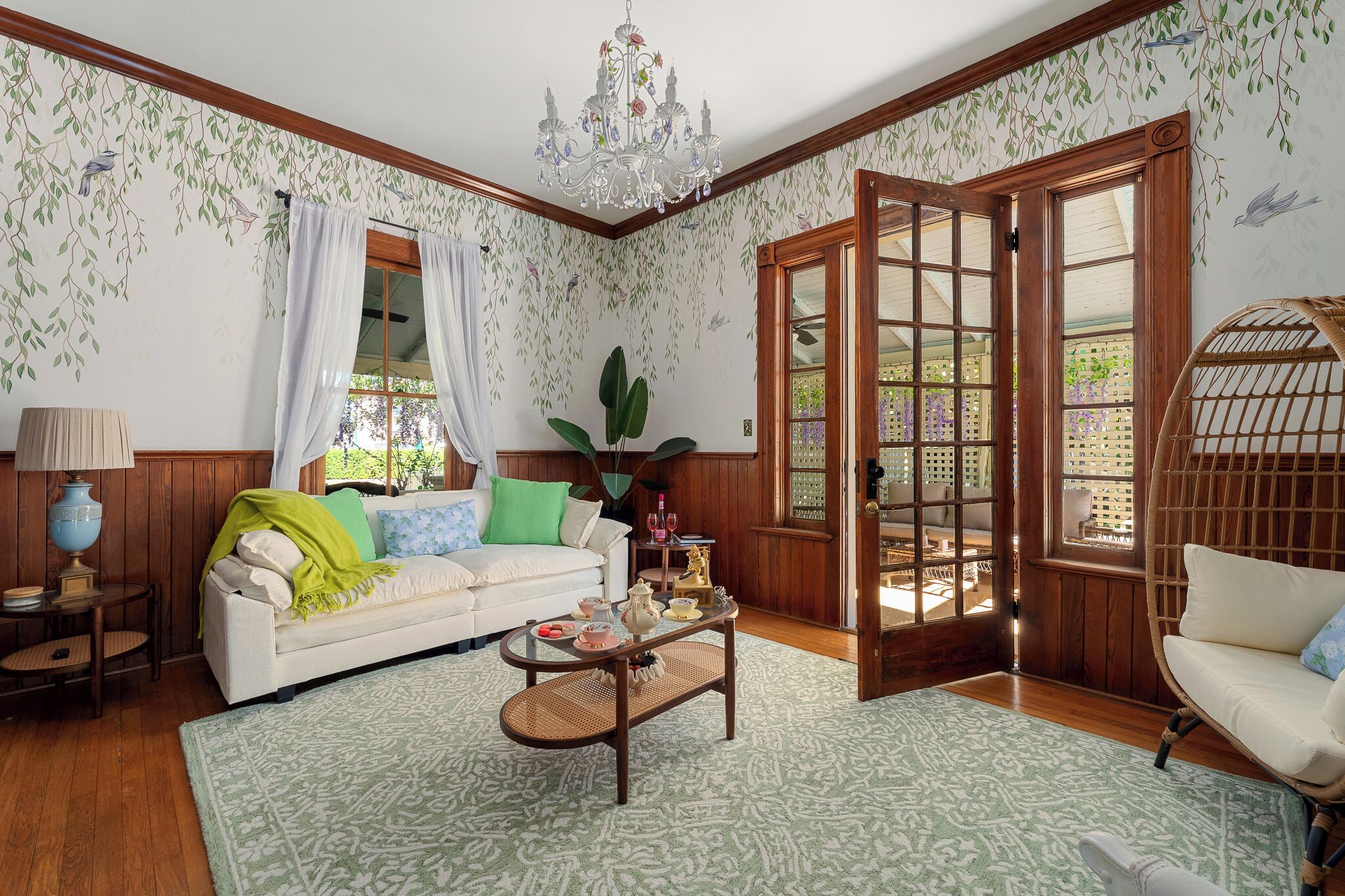 A cozy living room with a white sofa, a wicker chair, a coffee table, and a large window with curtains.