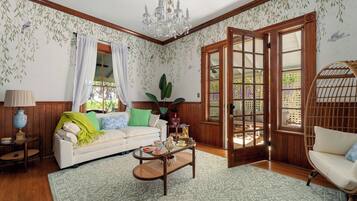 A cozy living room with a white sofa, a wicker chair, a coffee table, and a large window with curtains.