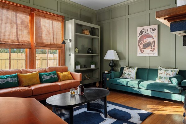 A living room with a green sofa, orange sofa, and a coffee table. There is a bookshelf, a lamp, and a poster on the wall.