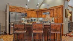 A kitchen with wooden cabinets, a stainless steel refrigerator, a microwave, and a gas stove. There are three bar stools and pendant lights above the island.